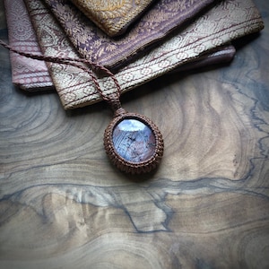 Op de afbeelding: Een ketting van bruin lederen koord met een geweven hanger met een blauwe en grijze edelsteen.