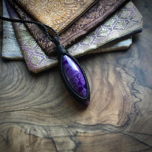 May include: A purple and white agate gemstone pendant with a black cord. The pendant is wrapped in black cord and has a teardrop shape.