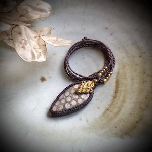 Op de afbeelding: Een handgemaakte ketting met een hanger van een patroonsteen en goudkleurige accenten. De hanger hangt aan een gevlochten koord. De ketting wordt gepresenteerd op een neutrale ondergrond.