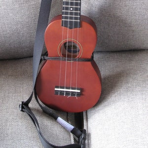 May include: A brown ukulele with a black strap, resting on a gray tweed fabric. The ukulele has the words "DIAMOND HEAD" on the headstock. The instrument has four strings and a round sound hole.