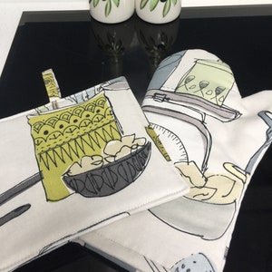 May include: Two white ceramic olive oil bottles with green olive branch designs and the words "olio" and "auto" on them.  Two white and grey kitchen pot holders with a black and white illustration of kitchen utensils and food.