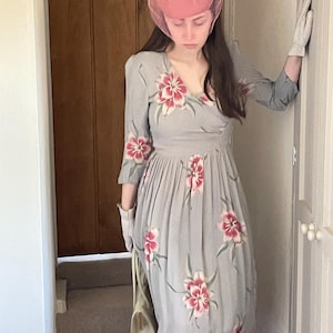May include: A woman wearing a grey floral dress with a pink hat and white gloves. She is holding a white handbag and standing on a carpeted staircase.