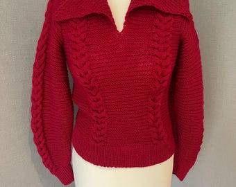 Op de afbeelding: Een rode gebreide trui met een brede kraag en een V-hals. De mouwen zijn lang en hebben een kabelgebreid ontwerp. De body van de trui heeft ook een kabelgebreid patroon. De trui wordt op een mannequin getoond.