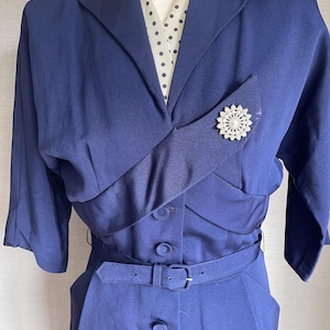 Könnte beinhalten: Ein marineblaues Vintage-Kleid mit maßgeschneidertem Design. Das Kleid hat einen Reverskragen, eine dekorative Brosche und einen passenden Gürtel. Das Kleid hat kurze Ärmel und eine Knopfleiste vorne. Eine weiße Bluse mit schwarzen Punkten ist darunter sichtbar.