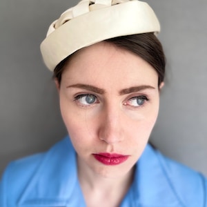 May include: Cream-colored woven hat with a rounded crown. The hat is worn by a person wearing a light blue jacket. The background is a solid gray.