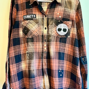 May include: A black and orange plaid flannel shirt with bleach splatters. The shirt has two patches, one that says "It is what it is" and another with a tree of life design.