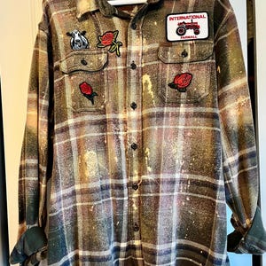 May include: A green, brown, and white plaid flannel shirt with bleach splatters. The shirt has several patches, including a cow, roses, and a red tractor with the text "INTERNATIONAL FARMALL".
