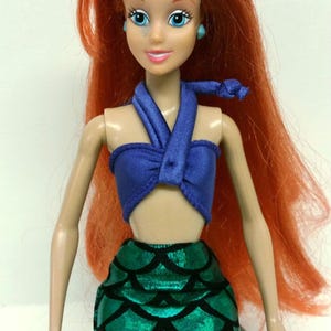 May include: A red-haired doll wearing a blue halter top and a green mermaid tail skirt. The doll has blue eyes and is wearing blue earrings.