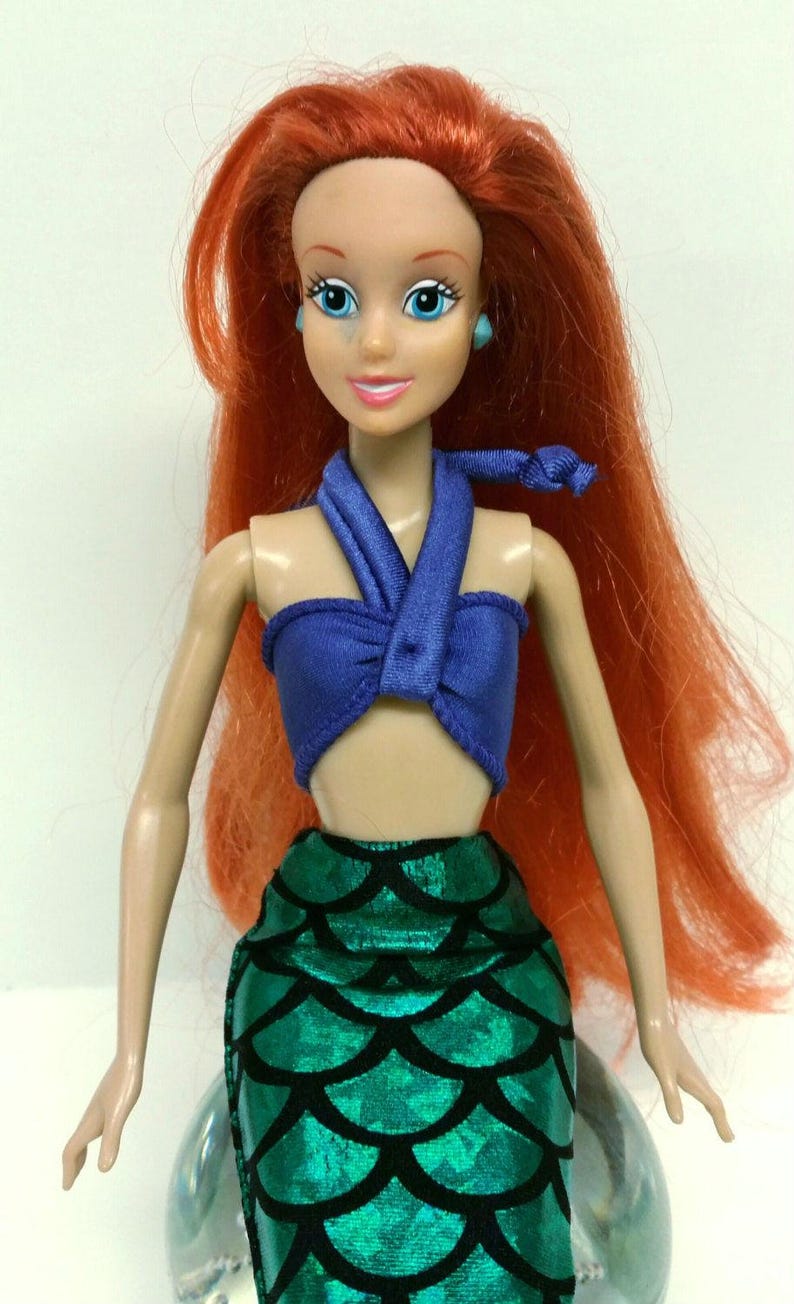 May include: A red-haired doll wearing a blue halter top and a green mermaid tail skirt. The doll has blue eyes and is wearing blue earrings.