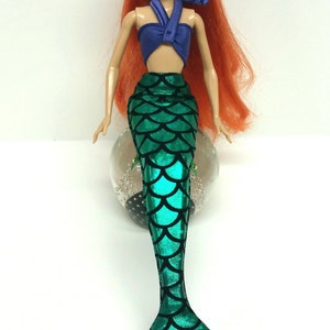 May include: A red-haired doll wearing a purple halter top and a green mermaid tail with a scale pattern.