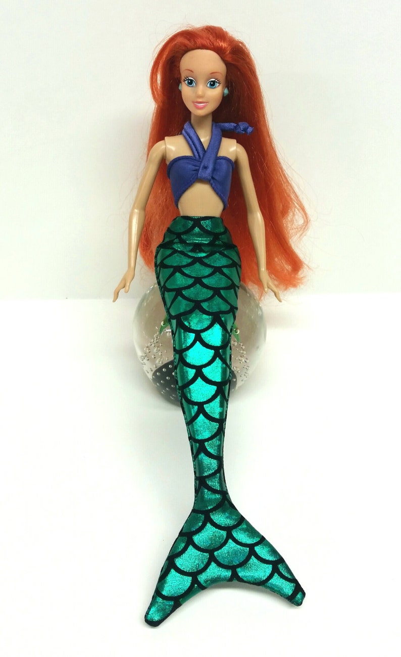 May include: A red-haired doll wearing a purple halter top and a green mermaid tail with a scale pattern.