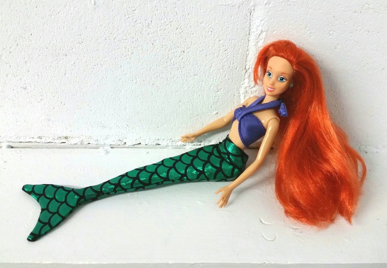 May include: A red-haired mermaid doll with a purple top and a green, glittery tail. The doll is lying on a white surface.