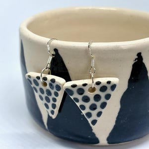May include: A pair of triangular ceramic earrings featuring a white base with black polka dots. The earrings are suspended from silver-toned metal hooks. Each triangular piece has a small hole at the top for the metal jump ring attachment.