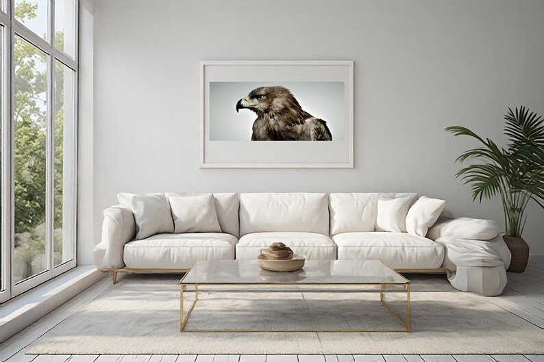 May include: A rectangular wall art print featuring a detailed profile portrait of a brown eagle with a sharp beak and intense gaze, displayed in a thin white frame centered on a plain light gray wall above a white sofa and glass coffee table.
