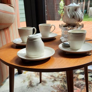 May include: A round wooden table with four white teacups and saucers, and a white teapot with floral accents. The table is set on a patterned rug, with a window and outdoor view in the background.