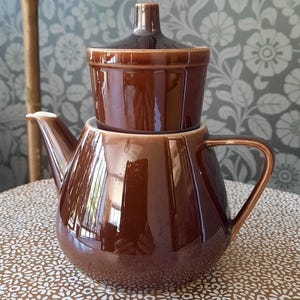 Op de afbeelding: Een bruine keramische theepot met een bijpassend deksel en een aparte, kleinere container die erop is gestapeld. De theepot heeft een gebogen lichaam, een tuit en een handvat. De set staat op een tafel met een tafelkleed met bloemenpatroon.