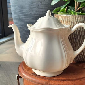 Può includere: Teiera in ceramica color crema con coperchio abbinato. La teiera ha un beccuccio e un manico curvi e poggia su un tavolino rotondo in legno. Lo sfondo comprende piante in vaso e una finestra.
