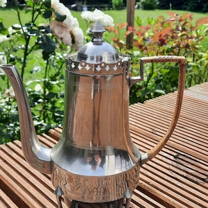 Pode incluir: Uma cafeteira de metal prateado com um corpo cilíndrico alto, uma tampa pontiaguda com uma borda recortada decorativa, um bico curvo e uma alça de vime trançado castanho claro. A base é apoiada por três pequenos pés de metal.