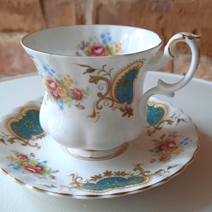 Puede incluir: Juego de taza y platillo blancos con detalles florales y dorados. La taza y el platillo presentan un diseño de cachemira azul y dorado. La taza tiene un asa curvada y se asienta sobre un platillo a juego, creando un elegante juego de té.