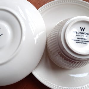 May include: White Wedgwood Windsor teacup, saucer, and plate set. The teacup has a ribbed design and a handle. The saucer and plate have a beaded edge. The bottom of the teacup and saucer have the Wedgwood logo and text.