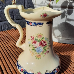 Op de afbeelding: Crèmekleurige keramische kan met een gebogen handvat en bloemenmotief. De kan heeft een boeket van rode, roze en blauwe bloemen. Donkerblauwe banden met bloemaccenten sieren de boven- en onderkant. Een decoratief stuk om te serveren.