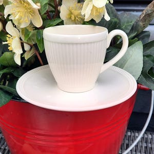 May include: A cream-colored teacup and saucer set with vertical ribbed detailing sits atop a red bucket. The bucket has a wooden handle and is filled with light yellow and green flowers. The scene evokes a sense of spring and outdoor decor.