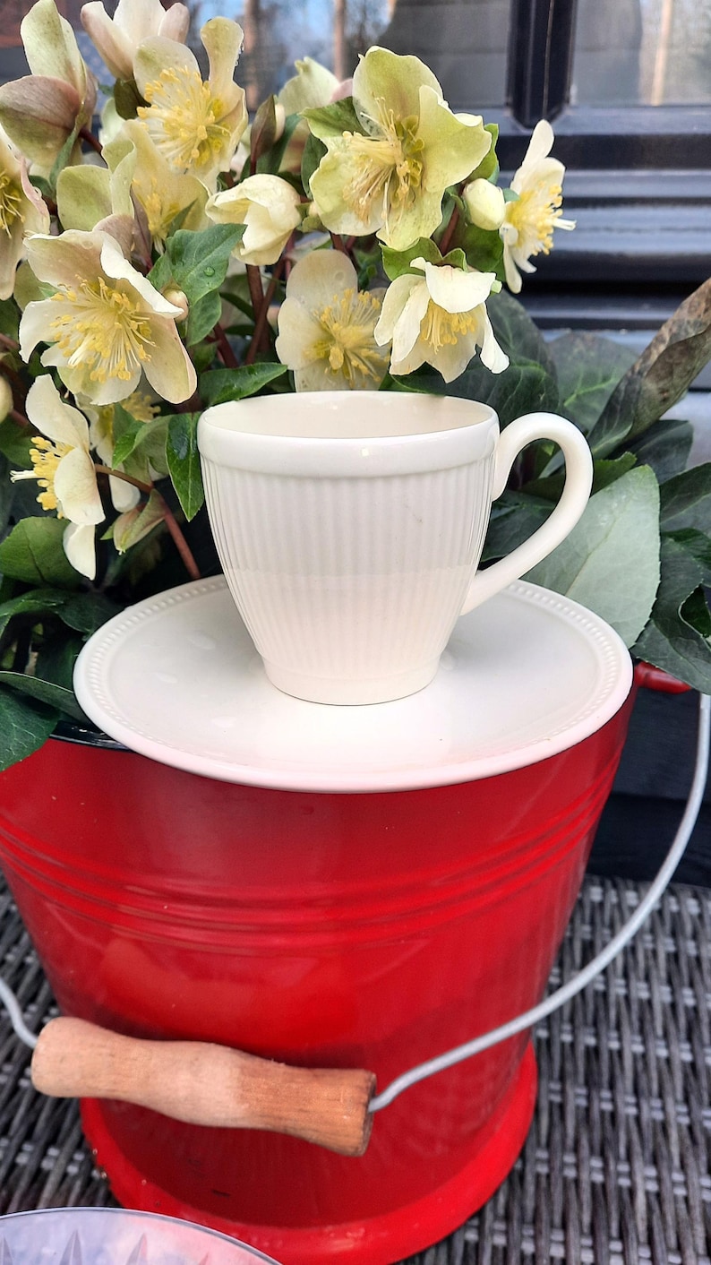 May include: A cream-colored teacup and saucer set with vertical ribbed detailing sits atop a red bucket. The bucket has a wooden handle and is filled with light yellow and green flowers. The scene evokes a sense of spring and outdoor decor.