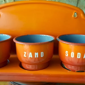 May include: Vintage orange metal kitchen organizer with three round containers. The containers have blue interiors and the word "SODA" or "ZAND" in white lettering. The organizer has a matching backboard.