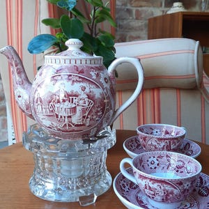 Può includere: Un servizio da tè vintage con una teiera, due tazze e piattini. La teiera e le tazze presentano un motivo a scena pastorale rosso e bianco. La teiera poggia su uno scaldavivande in vetro trasparente.