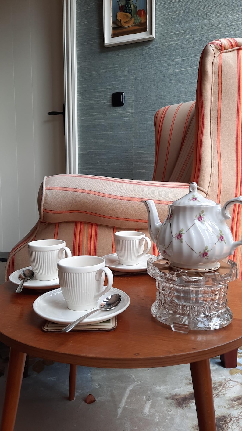 May include: A white porcelain tea set with a floral design, including a teapot, cups, saucers, and spoons, sits on a wooden table. The teapot rests on a glass trivet. The scene is set in a room with a striped armchair.