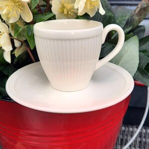May include: A cream-colored teacup and saucer set with vertical ribbed detailing, resting on a red bucket. The teacup has a curved handle. The background features yellow and green flowers.