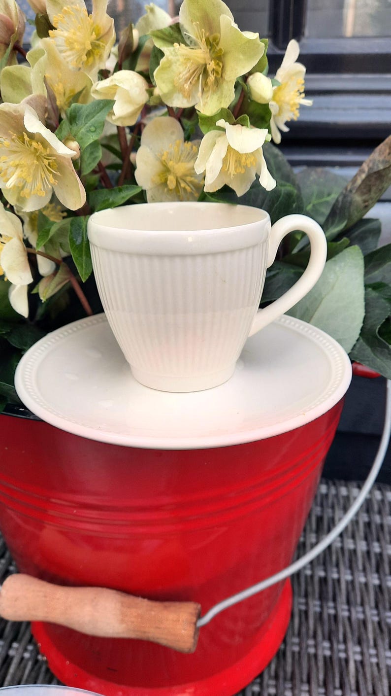 May include: A cream-colored teacup and saucer set with vertical ribbed detailing, resting on a red bucket. The teacup has a curved handle. The background features yellow and green flowers.