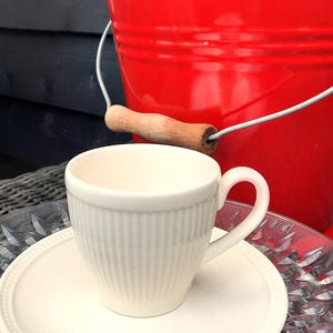 May include: A white teacup and saucer set with vertical ribbed detailing. The cup sits on a matching saucer, with a red bucket in the background. The bucket has a wooden handle and contains greenery and flowers.