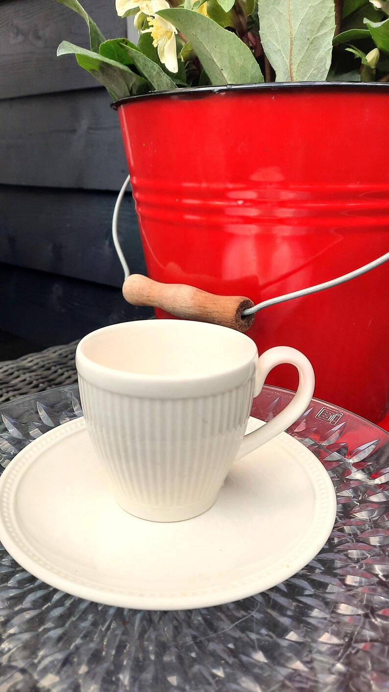 May include: A white teacup and saucer set with vertical ribbed detailing. The cup sits on a matching saucer, with a red bucket in the background. The bucket has a wooden handle and contains greenery and flowers.