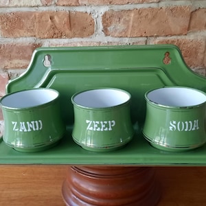 Può includere: Un organizer da cucina vintage in smalto verde con tre contenitori etichettati: ZAND, ZEEP e SODA. Il set include un vassoio e una piastra posteriore con fori per appenderlo. I contenitori hanno interni bianchi e finiture dorate.