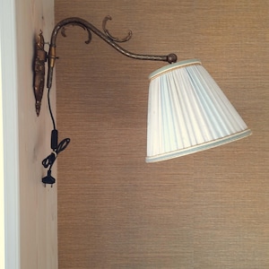 Könnte beinhalten: Eine Wandleuchte mit hellblauem, plissiertem Lampenschirm und einem dekorativen Arm in Bronzefarbe. Die Lampe ist an einer hellen Wand befestigt und hat ein schwarzes Netzkabel.