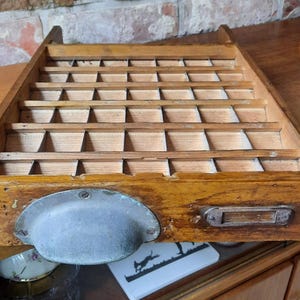 May include: A vintage wooden printer's tray with many small compartments. The tray has a metal handle and is made of light brown wood. The background includes a brick wall and a wooden cabinet.