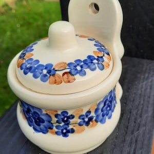 Może przedstawiać: Kremowy ceramiczny słoik z pokrywką, ozdobiony niebieskimi i brązowymi wzorami kwiatowymi. Słoik ma zaokrąglony kształt i mały uchwyt z tyłu do zawieszenia. Mały guziczek znajduje się na górze pokrywki.