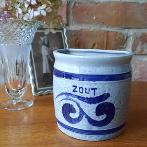 Op de afbeelding: Blauw-witte keramische pot met het woord "ZOUT" en een golfontwerp. De pot is cilindrisch en heeft een textuur. De achtergrond omvat een bakstenen muur, een vaas en een ingelijste foto.