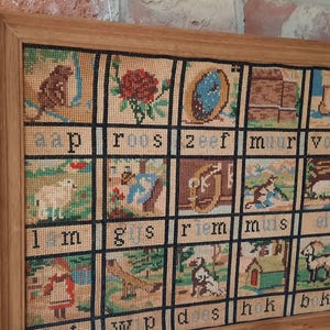 Può includere: Campione di ricamo vintage incorniciato con una griglia di quadrati colorati. Ogni quadrato presenta un'immagine diversa e una parola in testo nero. La cornice è in legno marrone chiaro. Lo sfondo è un muro di mattoni.