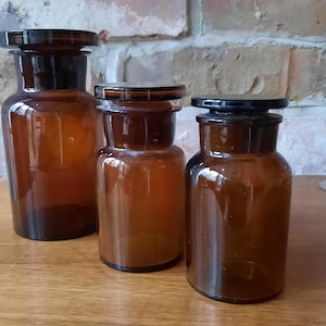 Op de afbeelding: Drie bruine glazen apothekerspotten met zwarte deksels. De potten hebben verschillende maten en een cilindrische vorm. De potten staan op een houten oppervlak tegen een bakstenen muur.