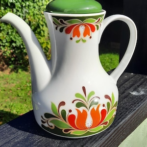 May include: White ceramic coffee pot with a green lid and handle. The pot is decorated with orange and red floral designs with green leaves. The pot has a long spout and is sitting on a dark surface.