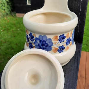 Op de afbeelding: Crèmekleurige keramische zoutpot met deksel, versierd met blauwe en oranje bloemmotieven. De zoutpot heeft een gebogen lichaam en een handvat om op te hangen. Het deksel is rond en ligt naast de pot.