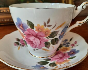 May include: White teacup and saucer set with a floral design featuring pink roses, blue and yellow flowers, and green leaves. The cup and saucer have gold trim. The handle of the teacup is also gold.