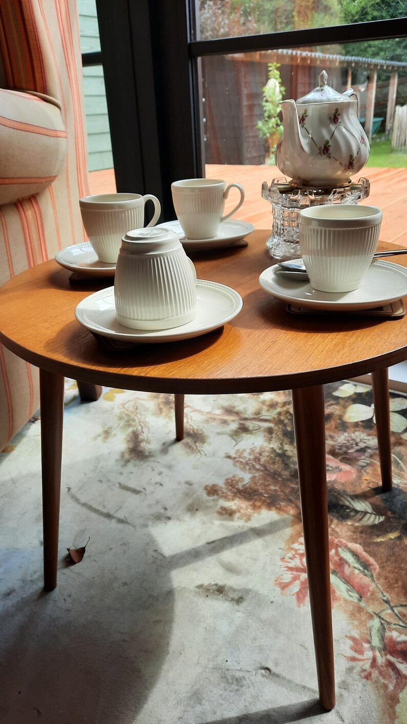 May include: A round wooden table with four white teacups and saucers, and a white teapot with floral accents. The table is set on a patterned rug, with a window and outdoor view in the background.