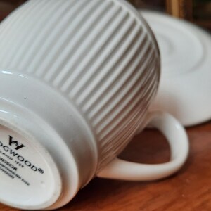 May include: White ceramic teacup and saucer set. The teacup has a ribbed design and a curved handle. The saucer is round and plain. The bottom of the teacup has the brand name "WEDGWOOD" and "WINDSOR" printed on it.