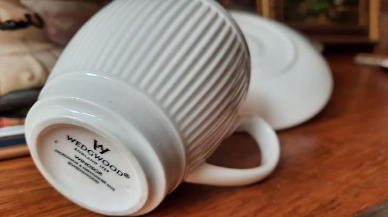 May include: White ceramic teacup and saucer set. The teacup has a ribbed design and a curved handle. The saucer is round and plain. The bottom of the teacup has the brand name "WEDGWOOD" and "WINDSOR" printed on it.