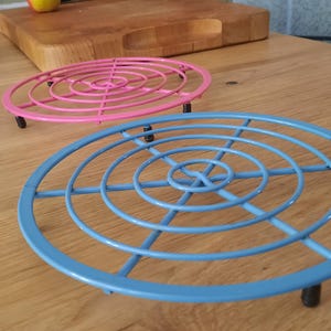 May include: Two circular metal trivets, one in light blue and one in pink, featuring a concentric ring and spoke design. Each trivet is supported by small black feet, providing a raised platform for kitchenware on a flat surface.