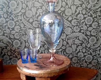 May include: A tall, iridescent blue glass decanter with a pink glass stopper and base, accompanied by one clear glass goblet and two small, vibrant blue shot glasses, all arranged on a circular wooden stand with a metallic rim.