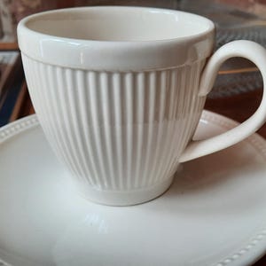 May include: A white ceramic teacup and saucer set. The teacup has vertical ribbed detailing and a curved handle. The saucer is round with a beaded edge. The set is placed on a wooden surface.